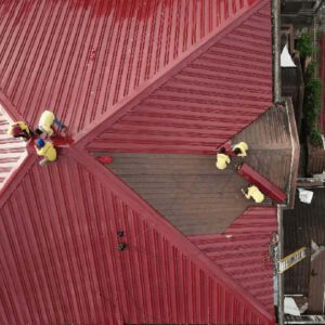 Workers repairing a red metal roof. This is another example of what you might need to plan for when you’re booking a commercial roof replacement.
