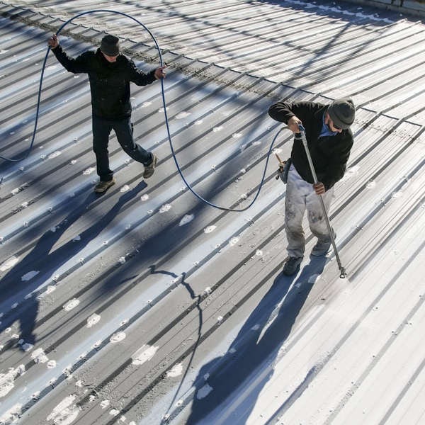two workers applying a protective coating on a metal commercial roof using spray equipment, demonstrating the restoration process to extend roof lifespan